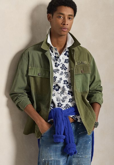 Πράσινο βαμβακερό μπουφάν με τσέπες, λευκό πόλο με λουλουδάτο σχέδιο, μπλε πλεκτό πουλόβερ δεμένο στη μέση και τζιν παντελόνι με φθορές.