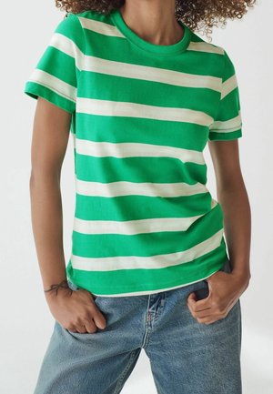 Person trägt grün-weiß horizontal gestreiftes Kurzarmshirt und blaue Jeans, Hände in den vorderen Taschen, mit sichtbarem lockigem Haar.