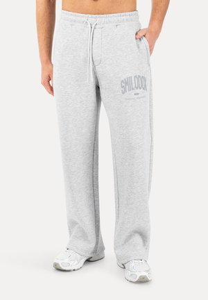 Person iført lysegrå joggingbukser med snøre og "SMILODOX" logo på låret, kombineret med hvide atletiksko.