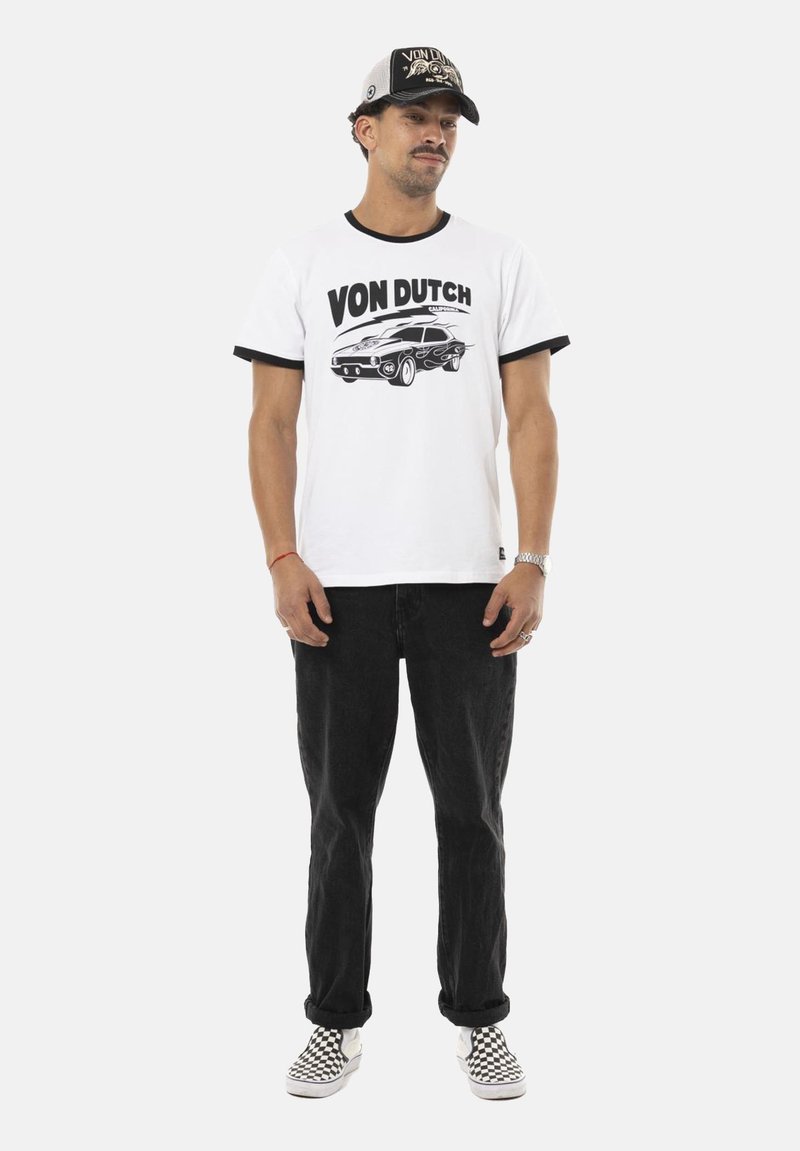 T-shirt blanc avec des accents noirs en anneau, portant le texte "VON DUTCH" et un graphique de voiture. Porté avec un jean noir et des chaussures à enfiler à carreaux.