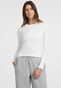Camicia bianca a maniche lunghe con scollatura tonda e un sottile dettaglio testuale sul petto, abbinata a pantaloni della tuta grigi con coulisse.