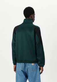 Πράσινο τζάκετ με ψηλό γιακά και μαύρες λεπτομέρειες, φτιαγμένο από λείο ύφασμα. Διαθέτει ραγλάν μανίκια και χαλαρή εφαρμογή, συνδυασμένο με μπλε denim.