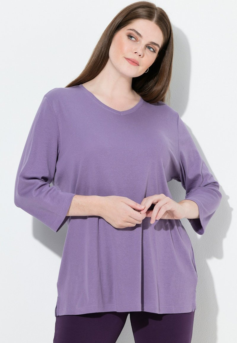 Top viola con scollo a V e maniche a tre quarti, realizzato in tessuto morbido che presenta una vestibilità rilassata e spacchi laterali per un comfort maggiore.