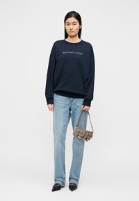 Marineblauer Kapuzenpullover mit "MICHAEL KORS" Stickerei, kombiniert mit hellblauen Jeans mit geradem Bein, grauer Handtasche und spitzen schwarzen Ballerinas.