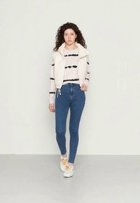 Colete branco acolchoado com capuz sobre um suéter creme com riscas pretas, combinado com jeans skinny azuis e ténis brancos com detalhes laranja.