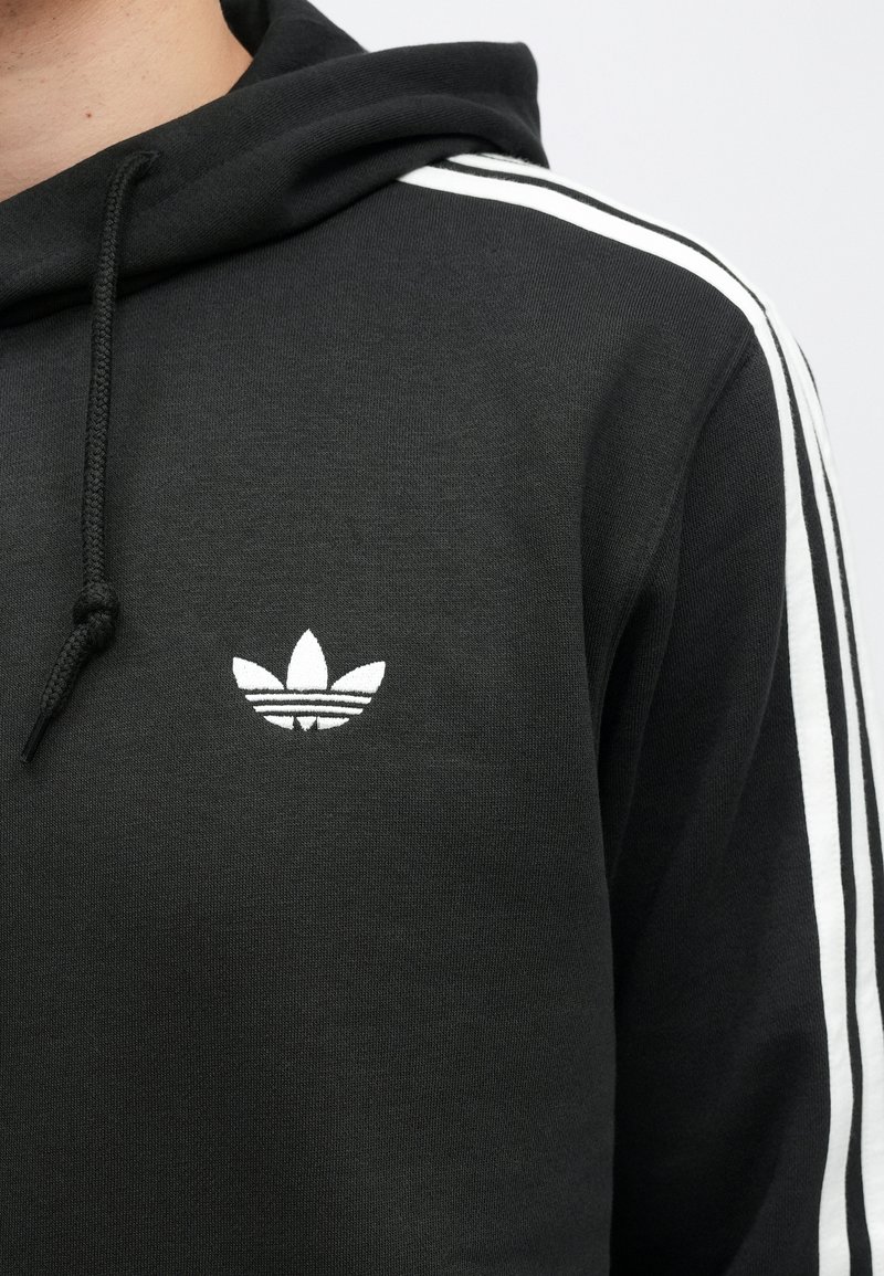 Felpa con cappuccio nera con logo trefoil Adidas bianco sul petto e tre strisce bianche che corrono sulla spalla e sulla manica.