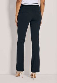Vrouw in fitted zwarte broek met achterzakken en beige hoge hakken, staande tegen een lichtgekleurde muur.