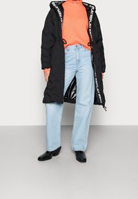 Černý péřový kabát s logovými akcenty, oranžový texturovaný rolák, světle modré džíny rovného střihu a černé vysoké boty.