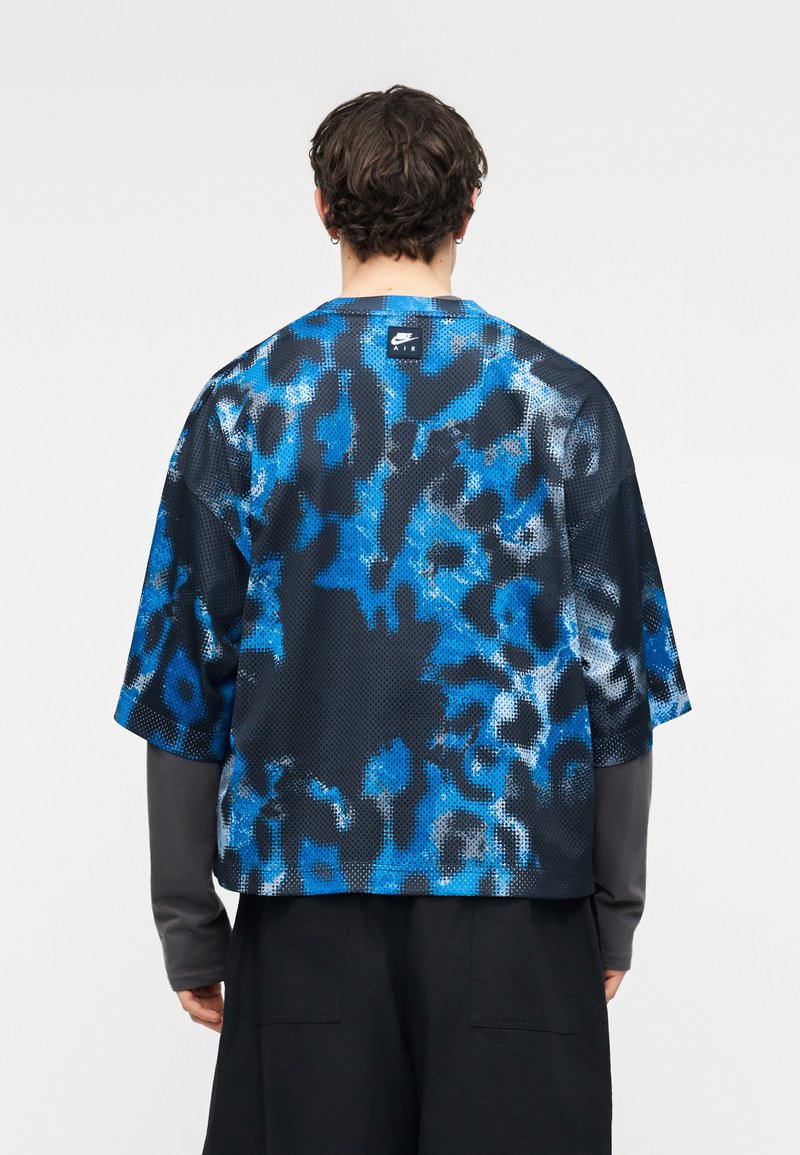 Person von hinten gesehen, trägt ein Nike Air Kurzarmshirt mit blau-schwarzem abstraktem Muster über einem langärmligen grauen Oberteil.