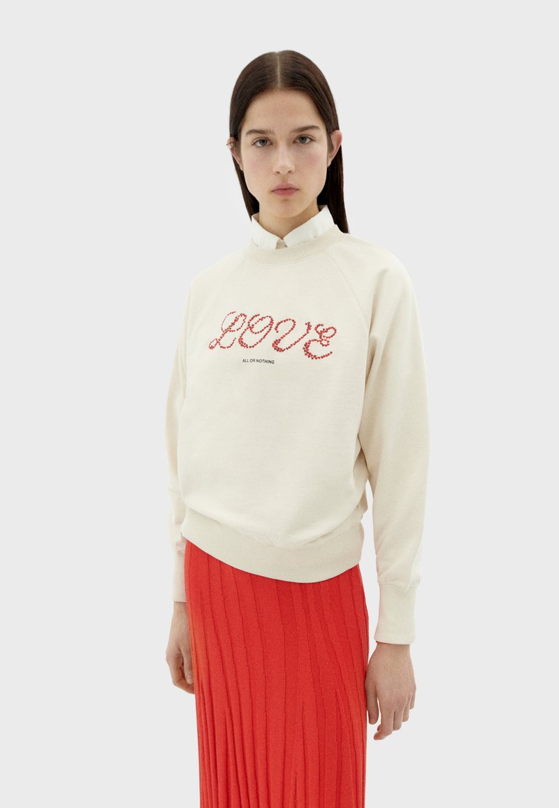 Cremfarbener Sweatshirt mit roter "LOVE" Stickerei, Rundhalsausschnitt und gerippten Bündchen, kombiniert mit einem roten, plissierten Rock.