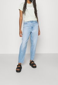 Maglietta verde chiaro con logo bianco, abbinata a jeans azzurri a vita alta e sandali neri. Taglio casual, materiale in cotone leggero.