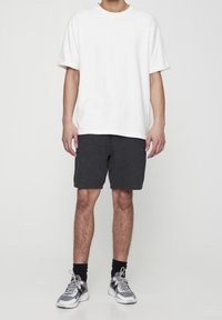 T-shirt blanche à manches courtes associée à un short gris foncé. Le modèle porte des baskets grises avec des accents noirs. Design simple, coupe décontractée.