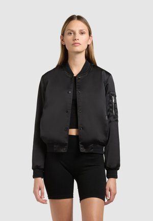 Woman wearing a black satin bomber jacket over a black crop top and black cycling shorts, standing against a plain light gray background.