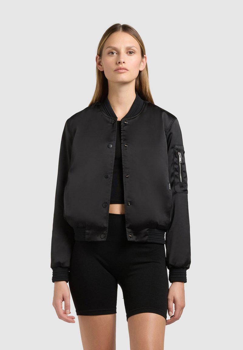 Femme portant une veste bomber en satin noir sur un crop top noir et un short de cyclisme noir, debout devant un fond gris clair uni.