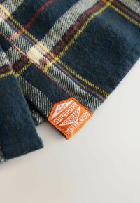 Marinblå och krämfärgad rutigt tyg med röda och gula accenter; har en texturerad yta. Orange vävd etikett visar "SUPERDRY STANDARD."