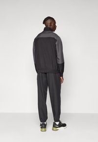 Schwarzer und grauer Trainingsanzug mit einer Zip-Jacke und elastischen Bündchen, kombiniert mit schwarzen Sneakers mit grünen Akzenten. Glatte Stofftextur.