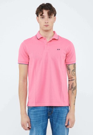 Giovane con capelli corti e ricci che indossa una polo rosa e jeans blu, mostra i tatuaggi sul braccio destro.