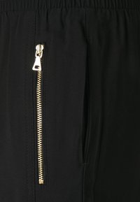 Schwarze Jogginghosen mit elastischem Taillenbund und einer Seitentasche, die einen goldenen Reißverschluss und kontrastierendes, strukturiertes Material aufweist.