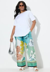 Haut blanc à manches courtes avec un col décoratif, associé à un pantalon floral à jambes larges dans des teintes de turquoise, orange et jaune. Sac à main argenté, sandales blanches.