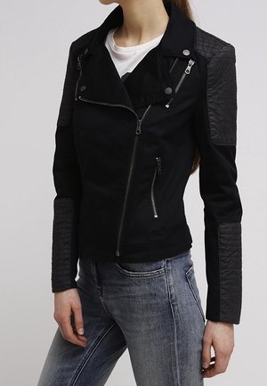 Femme portant une veste noire ajustée avec des patches matelassés sur les épaules et les avant-bras, associée à un jean bleu clair et une chemise blanche en dessous.