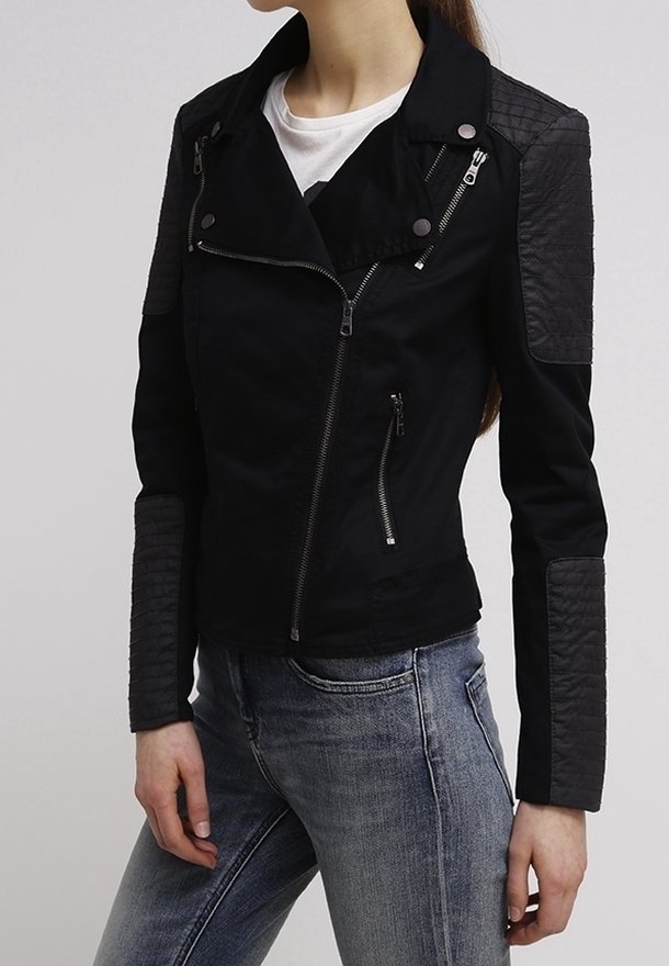 Femme portant une veste noire ajustée avec des patches matelassés sur les épaules et les avant-bras, associée à un jean bleu clair et une chemise blanche en dessous.