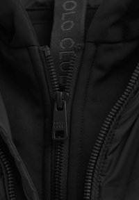 Närbild på en svart jacka med dragkedja, där "POLO CLUB" är ingraverat på dragkedjehänget, delvis uppdragen för att visa tyget under.