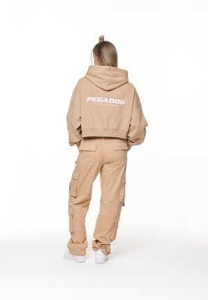 Personne portant un pantalon cargo beige et un sweat à capuche court avec le texte "PEGADOR" dans le dos, debout face à un fond blanc, de dos.