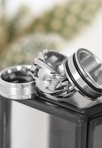 Drie metalen ringen: een gladde zilveren band, een gevlochten zilveren ontwerp en een band met zwarte accenten en een gepolijste afwerking.