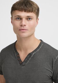Pelēks, īsām piedurknēm t-krekls ar V veida kakla izgriezumu un trim brūniem pogu akcentiem. Audumam ir mīksta tekstūra, ar izbalējušu dizainu ap kakla izgriezumu.