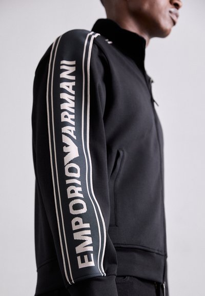 Musternukk musta tõmblukuga pesu, millel on kõrge kaelus, valge logo "EMPORIO ARMANI" varrukal ja kontrastsed triibud.