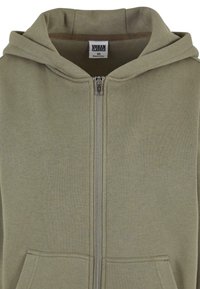 Sudadera con cremallera de color verde oliva, hecha de tela suave, con capucha, bolsillos frontales y una cremallera suave. La etiqueta dice "Urban Classics XS".