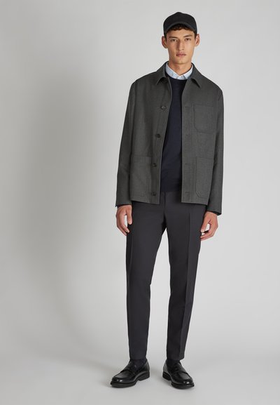 Giacca di lana grigia con colletto, due tasche frontali e chiusura con bottoni, abbinata a pantaloni scuri e scarpe nere. Texture liscia, design minimalista.