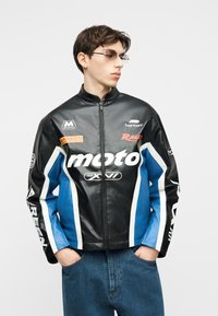 Jeune homme portant une veste de moto noire et bleue avec des rayures blanches et des logos, les mains dans les poches, regardant sur le côté.