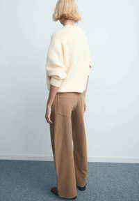 Pull crème tricoté avec épaules tombantes, associé à un pantalon marron à jambes larges avec poches arrière, porté avec des chaussures noires.