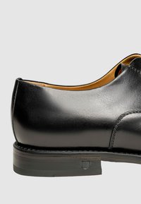 Schwarzer Lederschuh mit glänzender Oberfläche, runder Zehenpartie und einer dezenten Naht entlang der Nahtstelle. Mit niedrigem, gestapeltem Absatz.