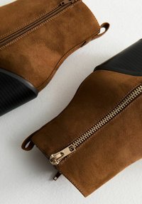 Brązowe zamszowe botki za kostkę z gładką czarną gumową podstawą, z złotymi zamkami błyskawicznymi i teksturowanymi akcentami. Projekt zawiera zaokrąglone czubki.