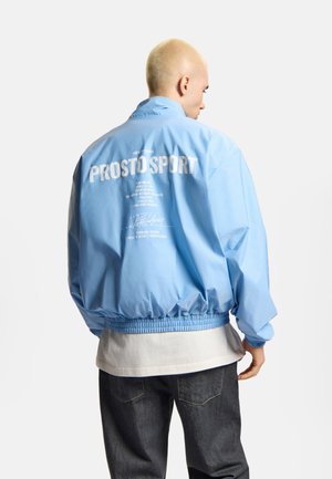 Osoba o krótkich blond włosach, ubrana w jasnoniebieską kurtkę wiatrówkę z białym napisem "PROSTO SPORT" na plecach, w połączeniu z ciemnymi dżinsami.