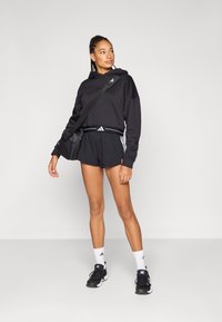 Zwarte cropped hoodie met logo, gecombineerd met zwarte shorts met witte strepen. Zwarte sneakers en witte sokken maken de sportieve look compleet.