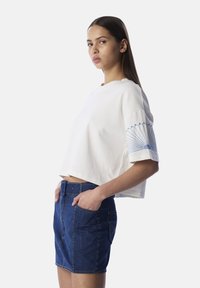 Wit cropped t-shirt met blauwe fanachtige borduursel op de linkermouw, gecombineerd met een donkerblauwe denim rok met voorzakken.
