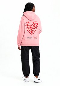 Felpa con cappuccio rosa con motivo a cuori rossi e testo "SWEET heart". Abbinata a pantaloni cargo neri e scarpe da ginnastica bianche. Tessuto morbido.