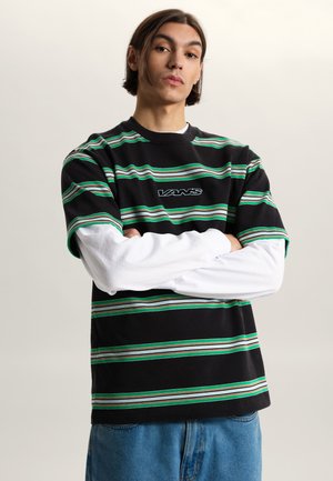 Junger Mann mit mittellangen Haaren trägt ein schwarz-grün-weiß gestreiftes Vans-Shirt über weißen Langarmärmeln und steht mit verschränkten Armen da.