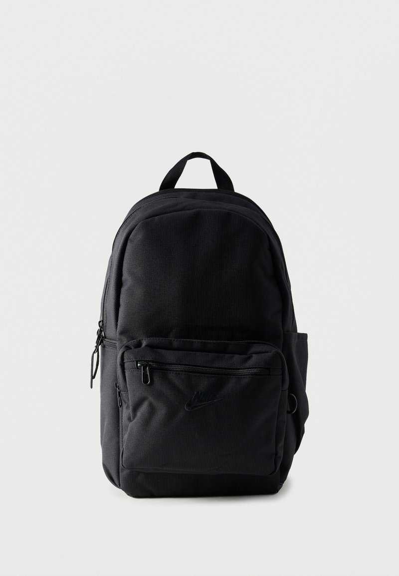 Sac à dos en tissu noir avec plusieurs compartiments à fermeture éclair et une poignée supérieure, posé debout sur un fond clair uni.