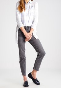 Lys grå cardigan over en blå og hvid ternet skjorte, parret med grå jeans og sorte loafers. Jeansene har en flosset kant.