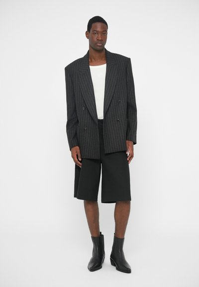 Dwurzędowa marynarka w paski w ciemnoszarym kolorze noszona na białej koszuli, w połączeniu z czarnymi szortami do kolan i czarnymi botkami za kostkę.
