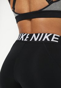 Leggings de sport noirs avec une large ceinture blanche portant la marque "NIKE". La texture est lisse avec une coupe ajustée. Le haut est un brassière de sport noire et grise.