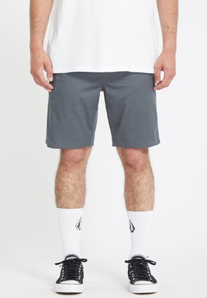Homme portant un short gris, des chaussettes blanches avec un logo, des baskets noires et un t-shirt blanc, debout devant un fond blanc uni.