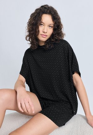 Tânără cu păr creț, stând jos, purtând un top negru lejer cu mâneci scurte și pantaloni scurți asortați, cu un model, pe un fundal simplu.