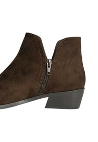 Bota de tobillo de ante marrón con cremallera lateral, tacón bajo y línea superior curva. Presenta una textura suave y detalles de costura mínimos.