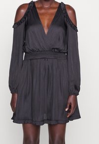 Svart klänning med djup V-ringning, kall axeldesign, långa ärmar, volangdetaljer vid axlarna och nederkant, samt en smockad midja. Slät textur.