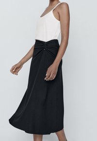 Gonna nera midi con dettaglio arricciato in vita e bottoni decorativi. Indossata con un top bianco senza maniche, caratterizzato da un tessuto liscio e un design fluido.